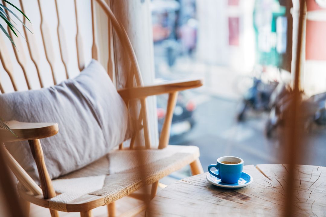 Ein Holzsessel mit Kissen am Fenster, auf dem Tisch eine blaue Espressotasse