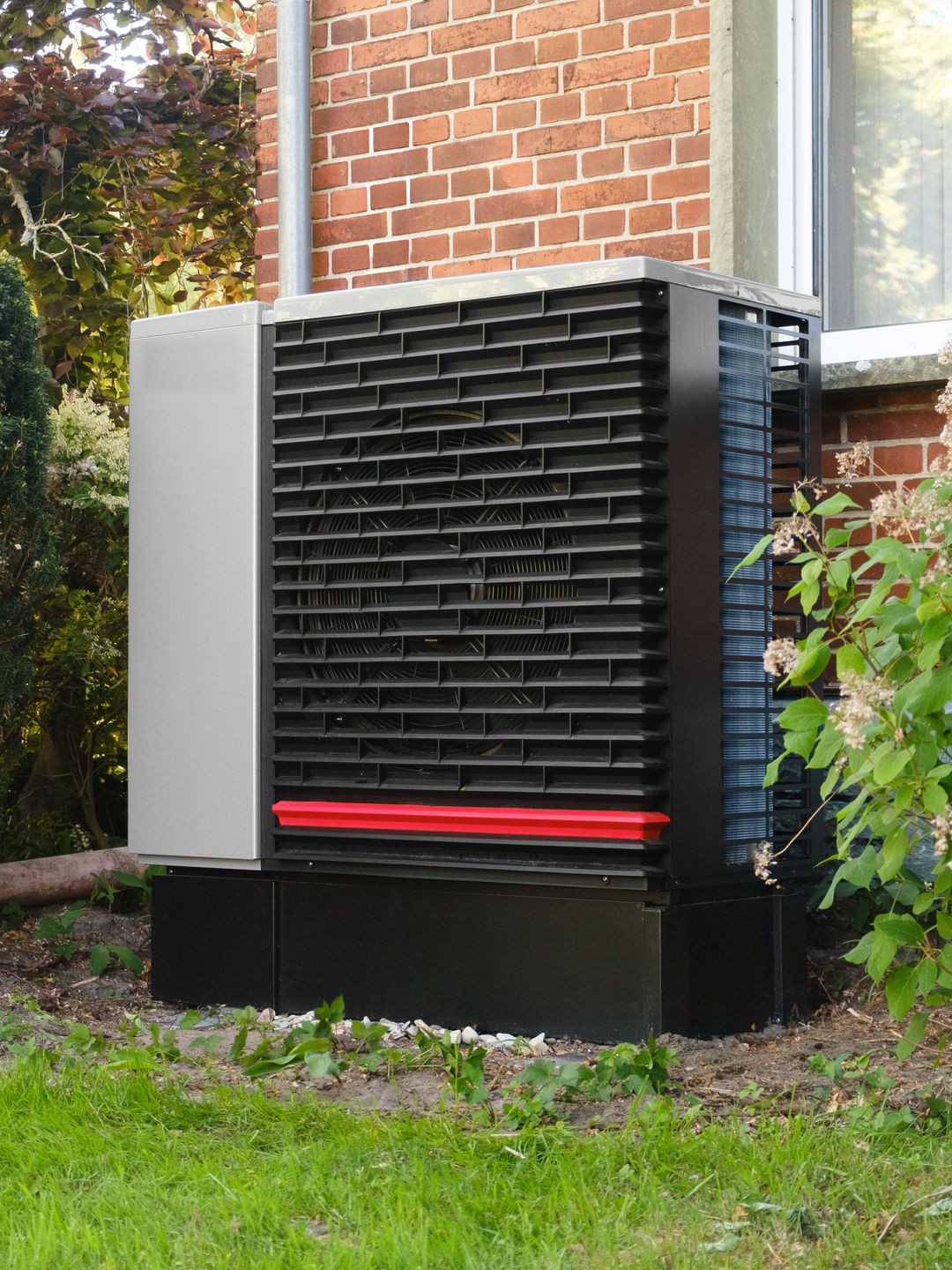 Eine Wärmepumpe mit rotem Streifen vor einem Klinkerhaus, zwischen Hecke und Gartenstrauch