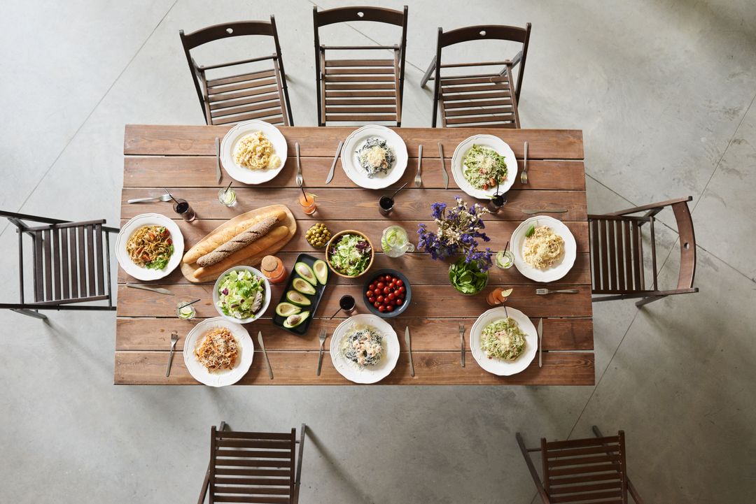 Eine lange Holztafel von oben, mit Tellern, Brot, Salaten und Blumen für eine ganze Runde