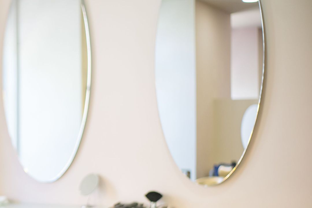 Eine Friseurin schneidet einer Kundin die Haare, an der hellen Wand hängen zwei ovale Spiegel, links fällt Licht durchs Fenster