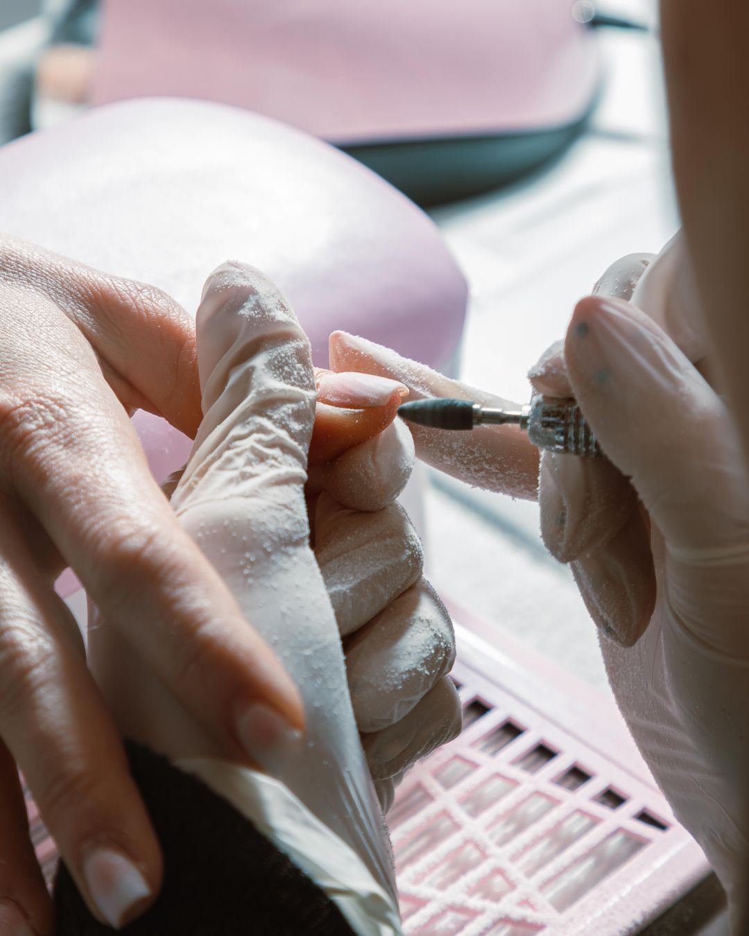 Hände in Handschuhen arbeiten mit einem feinen Fräser an der Nagelhaut, darunter die Absaugung